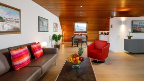 Un salon moderne avec un canapé en cuir marron, un fauteuil rouge et une table basse en verre sur laquelle sont disposés des fruits. On aperçoit à l’arrière-plan un coin repas avec une table et des chaises.