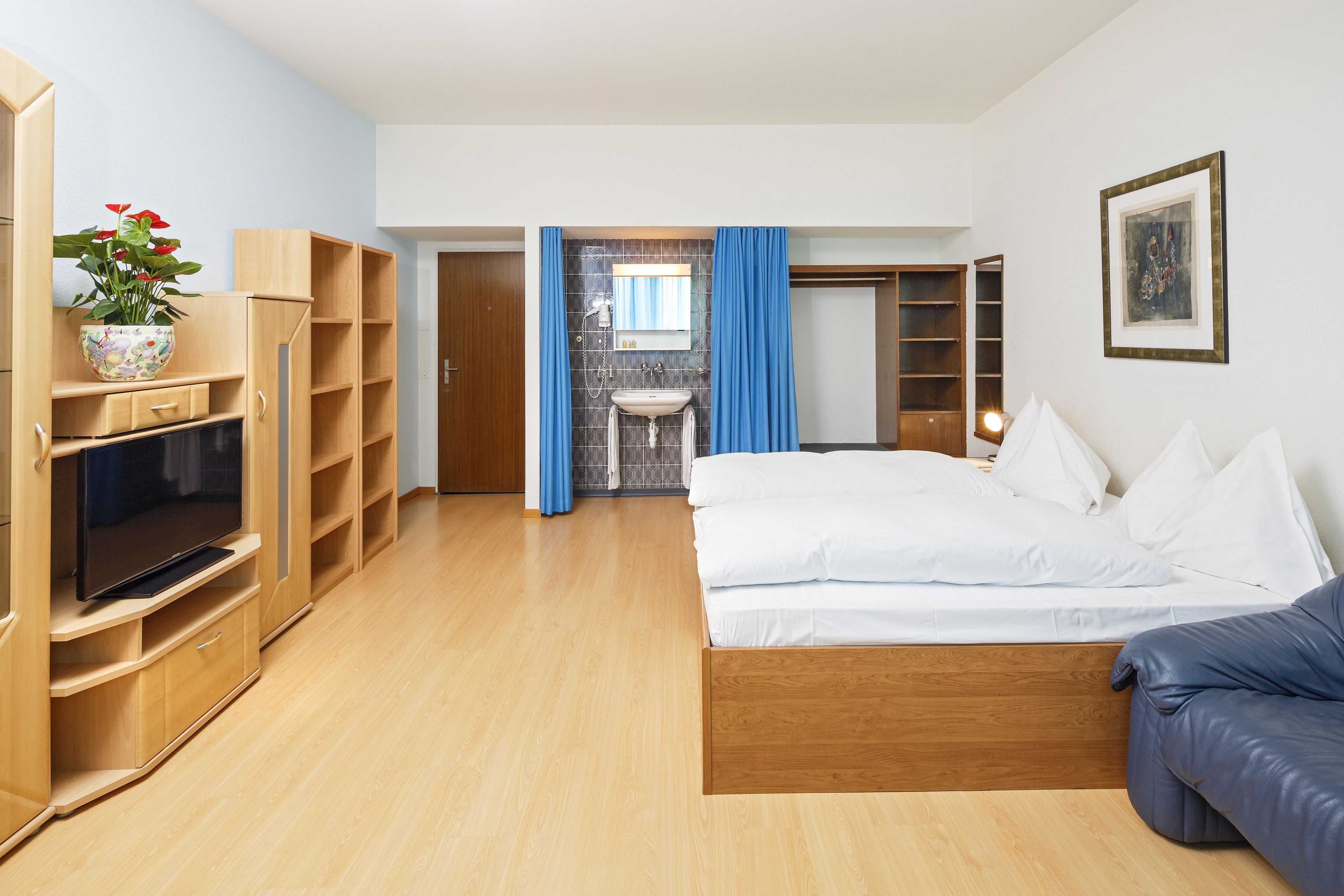 A modern hotel room with a wooden bed, a TV, a bathroom with a sink and mirror, and a blue curtain.