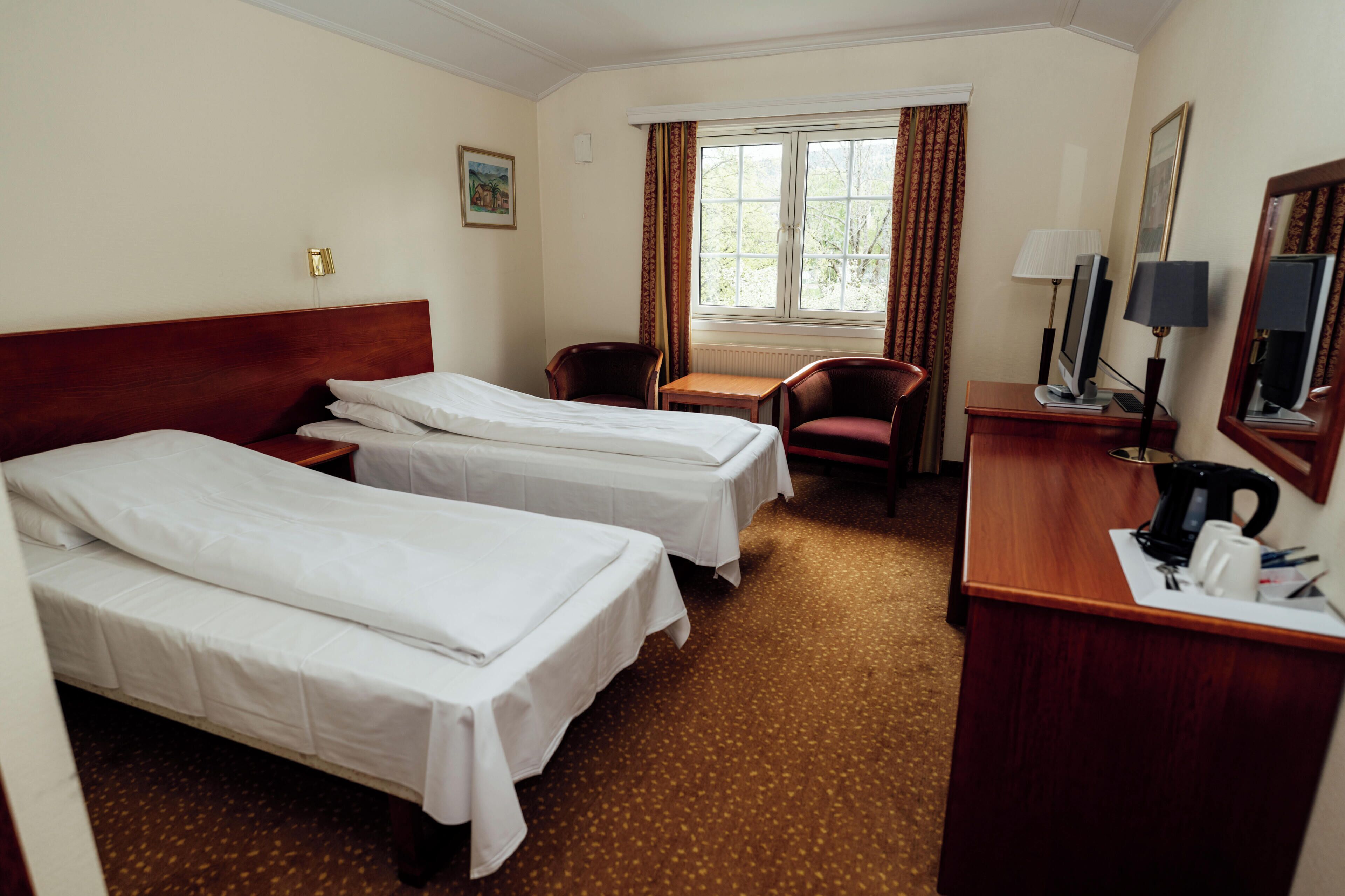 A hotel room with two single beds, a desk with a computer, a chair, and a window with curtains.