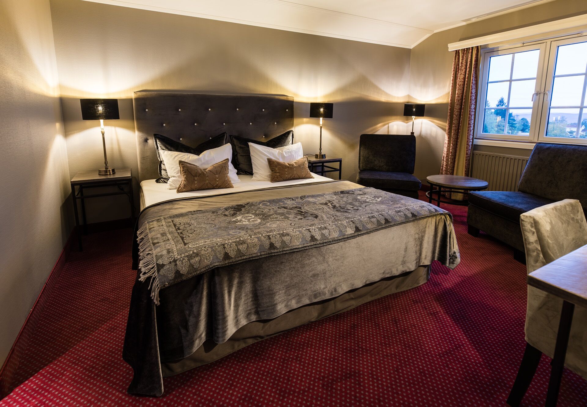 Ein Hotelzimmer mit einem großen Bett, zwei Nachttischen mit Lampen, einem kleinen runden Tisch, einer Couch und einem Fenster mit Vorhängen.