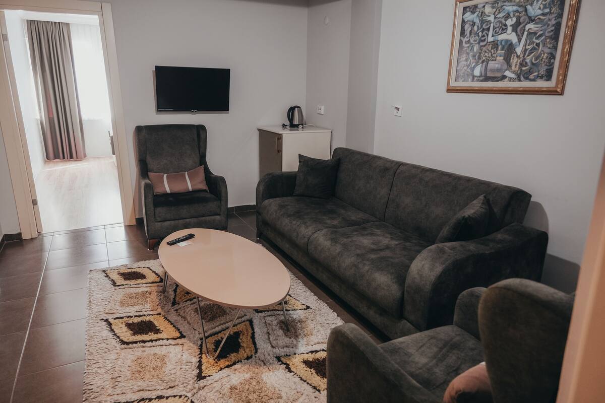 A living room with a grey sofa, a small round coffee table, a grey armchair, a TV on the wall, and a painting.
