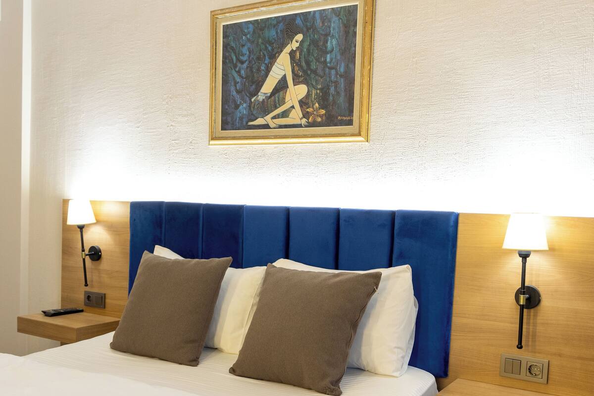 A hotel room with a large bed, two bedside tables, a blue headboard, and a framed picture on the wall.