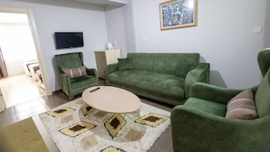 A living room with a green sofa, a matching armchair, a small table, and a TV on the wall.