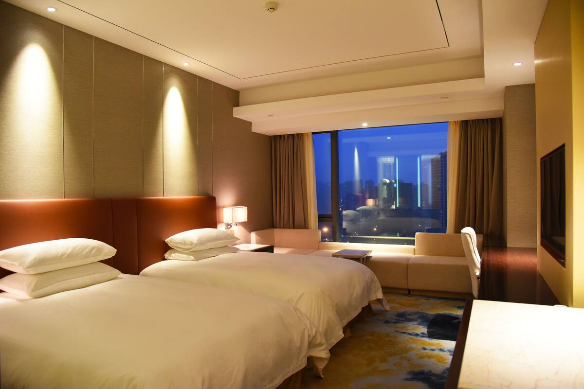 A hotel room with two beds, a sofa, a desk, and a large window with a city view.