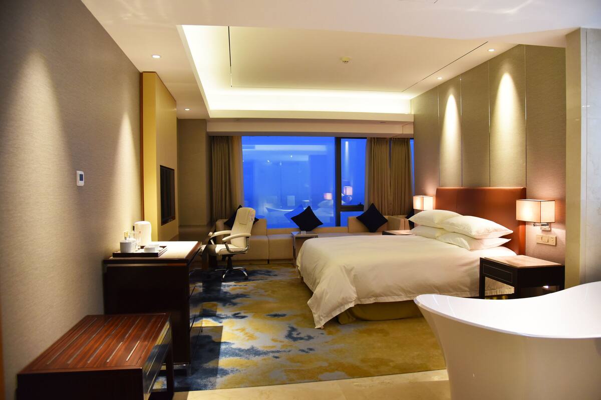A hotel room with a bed, a desk, a chair, a desk lamp, a bedside table, a nightstand, a bathroom with a bathtub, and a window with curtains.