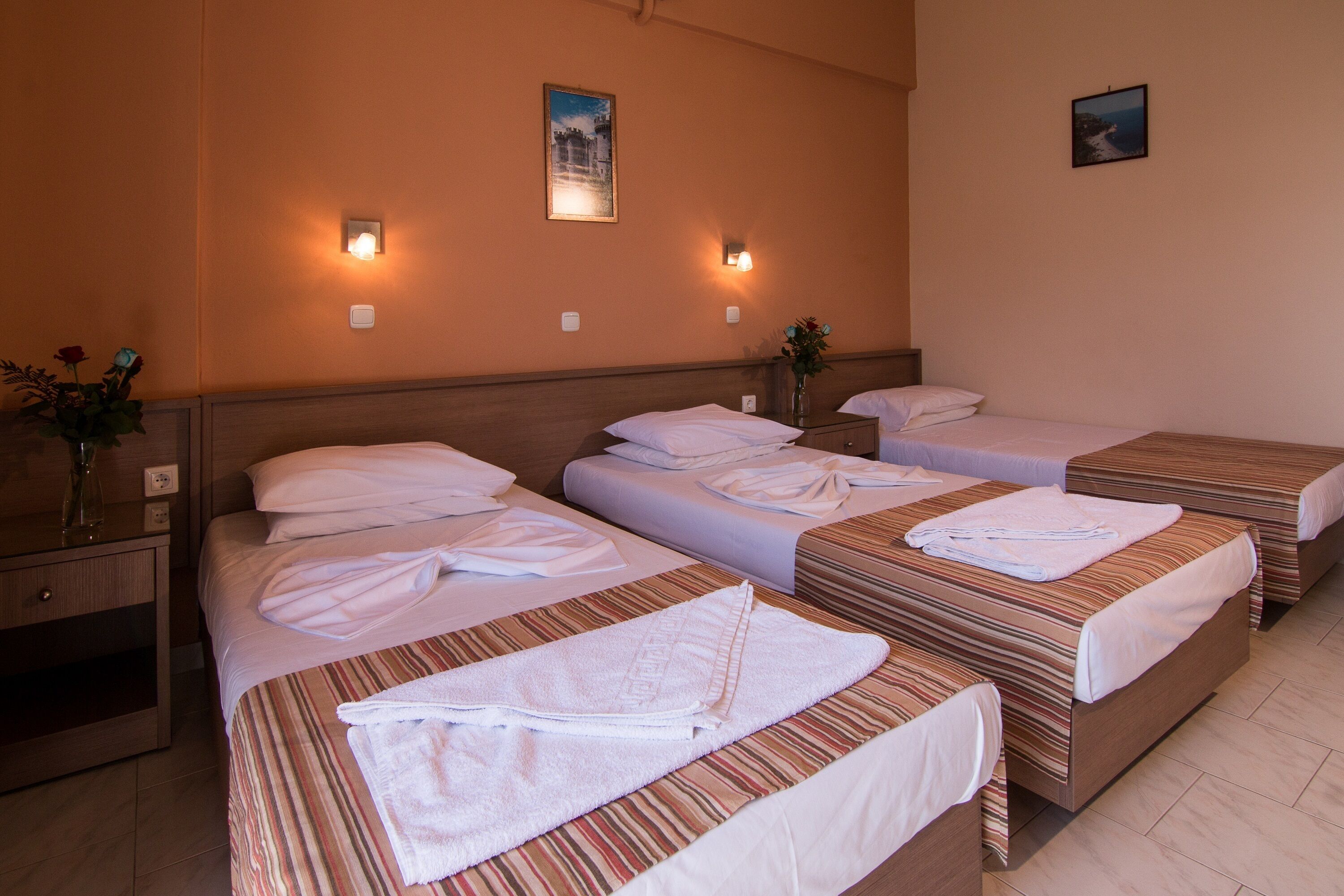A hotel room with three beds, each with a striped bedspread and white linens. There are wall-mounted lights, a vase with flowers on the nightstand, and a framed picture on the wall.