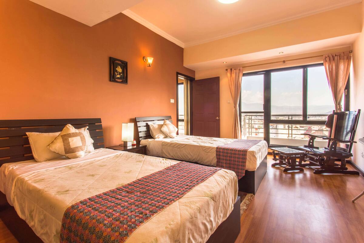 Habitación de hotel con dos camas, un escritorio con silla, un ventanal con vistas y un televisor montado en la pared.