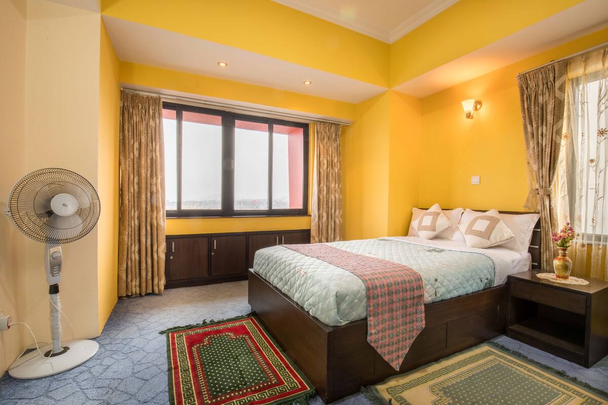 Un dormitorio con cama, mesita de noche, ventana con cortinas, un ventilador de pie y una alfombra estampada.