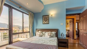 Une chambre avec un grand lit, une table de chevet, une commode en bois et une vue sur la ville grâce à de grandes fenêtres.