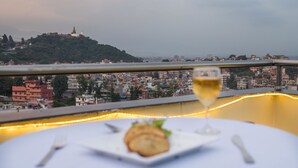 Une table à manger avec une assiette de nourriture et un verre de vin, donnant sur un paysage urbain avec des bâtiments et une colline surmontée d’une pagode.