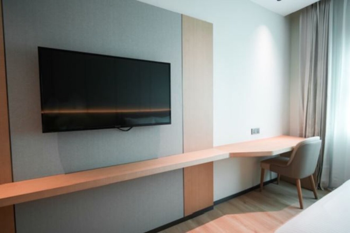 A modern hotel room with a flat-screen TV mounted on the wall, a wooden desk, a chair, and a bed.
