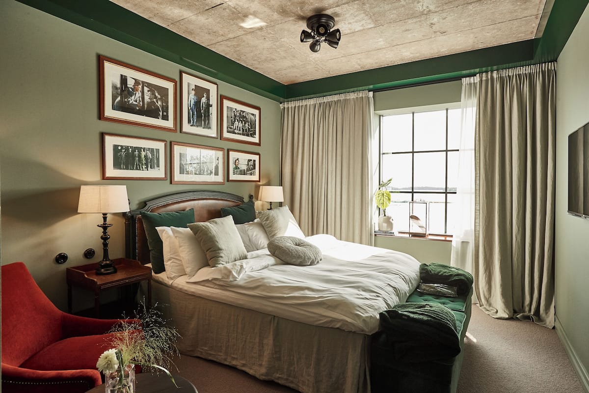 Un dormitorio con cama, mesita de noche, silla, ventana con cortinas e imágenes enmarcadas en la pared.