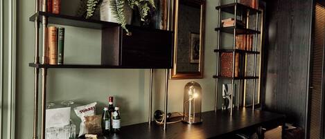 A bar area with a wooden counter, shelves with books and decorative items, a hanging plant, and a pendant light.