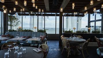Un ristorante con tavoli e sedie, ampie finestre vista sull'acqua e lampade a sospensione.