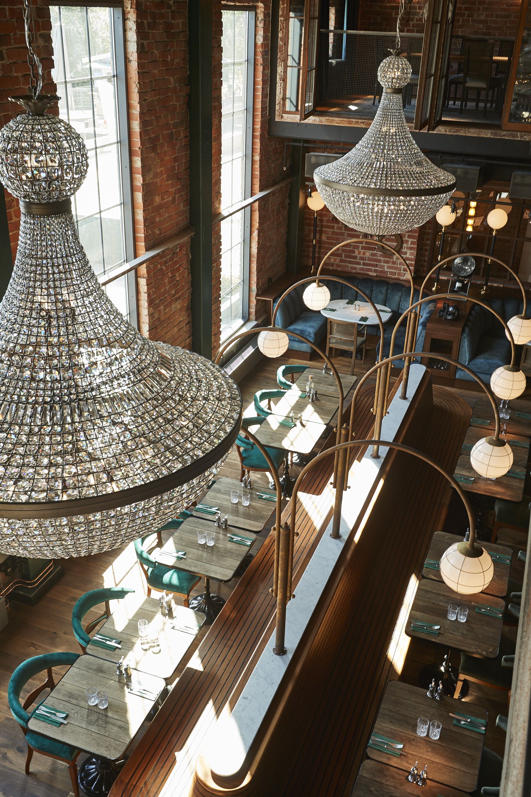 A restaurant with high ceilings, exposed brick walls, and multiple chandeliers. Tables are arranged in a linear fashion with green upholstered chairs.
