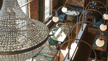 A restaurant with high ceilings, exposed brick walls, and multiple chandeliers. Tables are arranged in a linear fashion with green upholstered chairs.