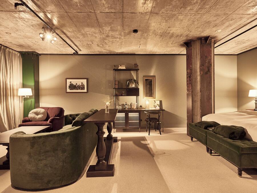 Habitación de hotel con una cama grande, un sofá verde, una mesa de centro de madera, un escritorio con una silla y una estantería.