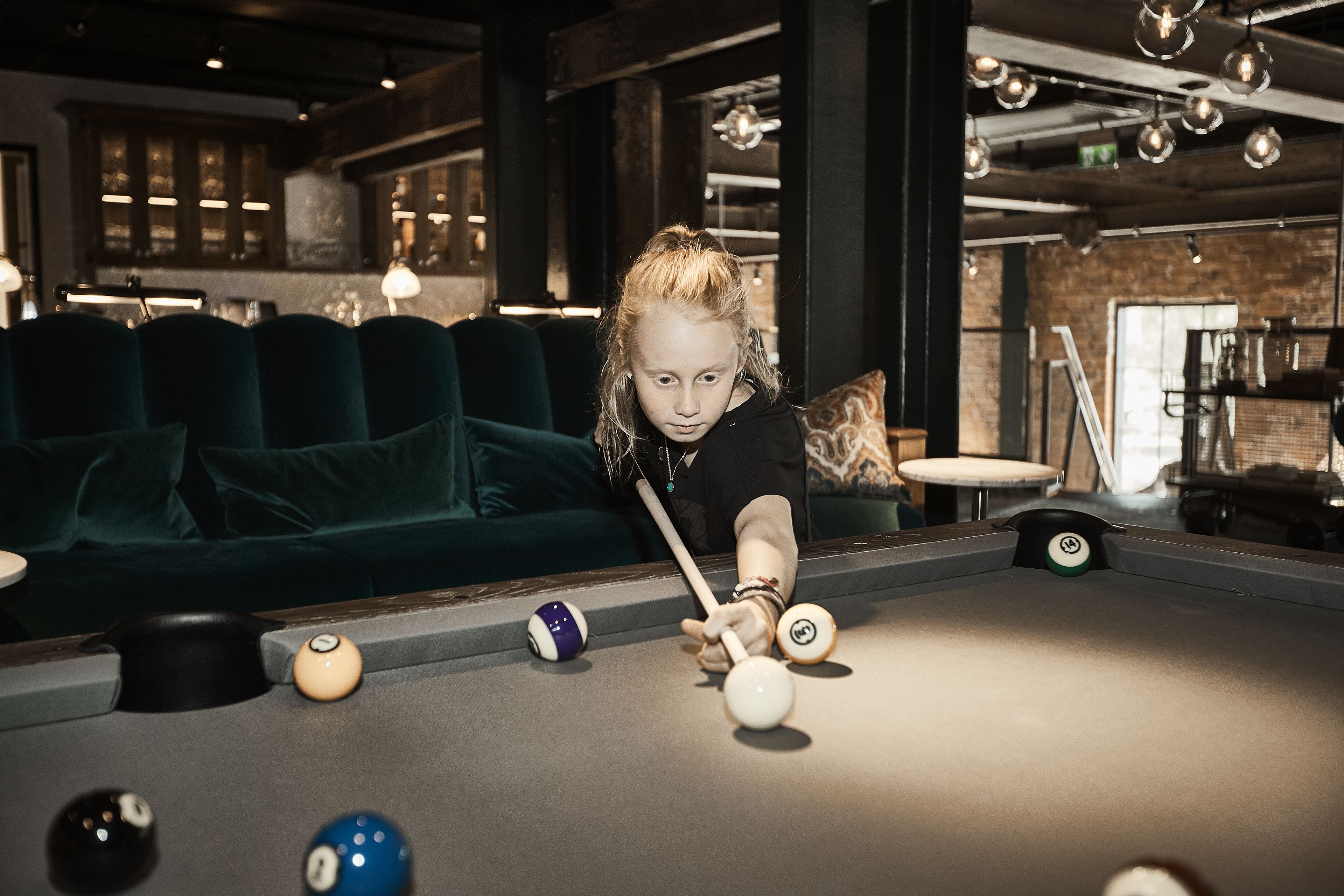 Une table de billard avec des billes et une queue, un canapé vert et un coin bar à l’arrière-plan.