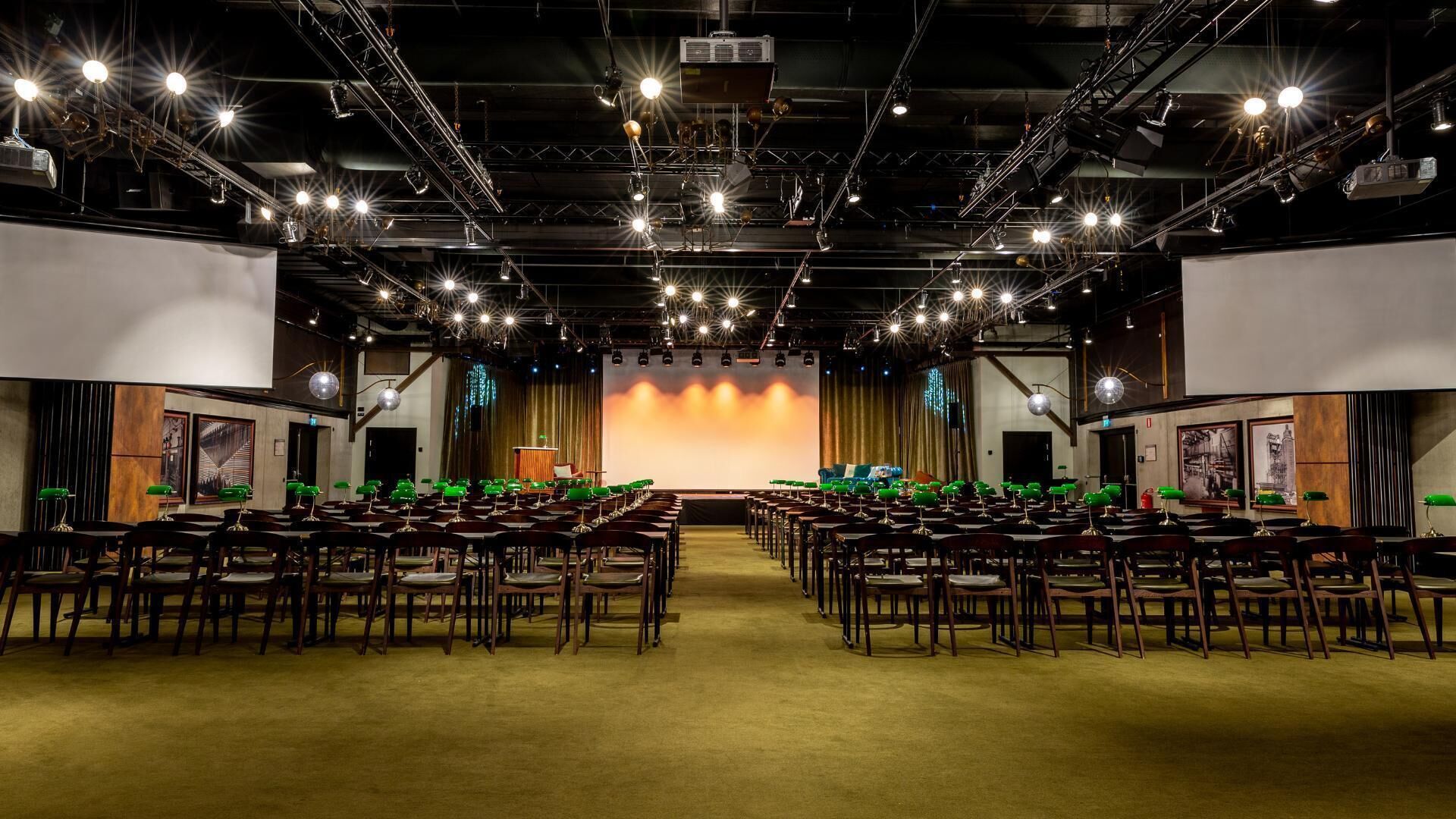 Un salón de conferencias con filas de sillas de madera, un escenario con un telón de fondo e iluminación colgante.