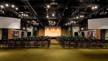 Un salón de conferencias con filas de sillas de madera, un escenario con un telón de fondo e iluminación colgante.