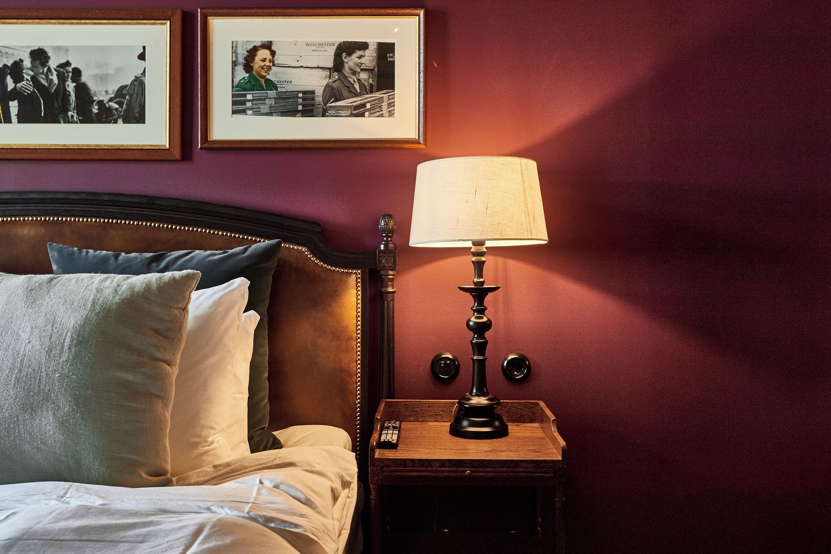 Un lit bien fait, avec des oreillers, une lampe de chevet, une télécommande et deux photographies encadrées accrochées au mur.