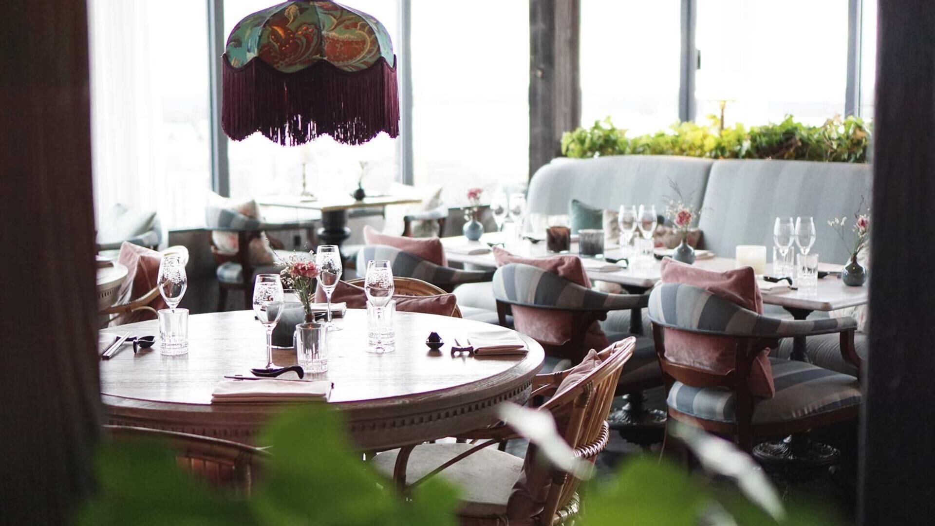 Un restaurante con mesas redondas y sillas acolchadas, ventanas amplias y una lámpara decorativa colgante.