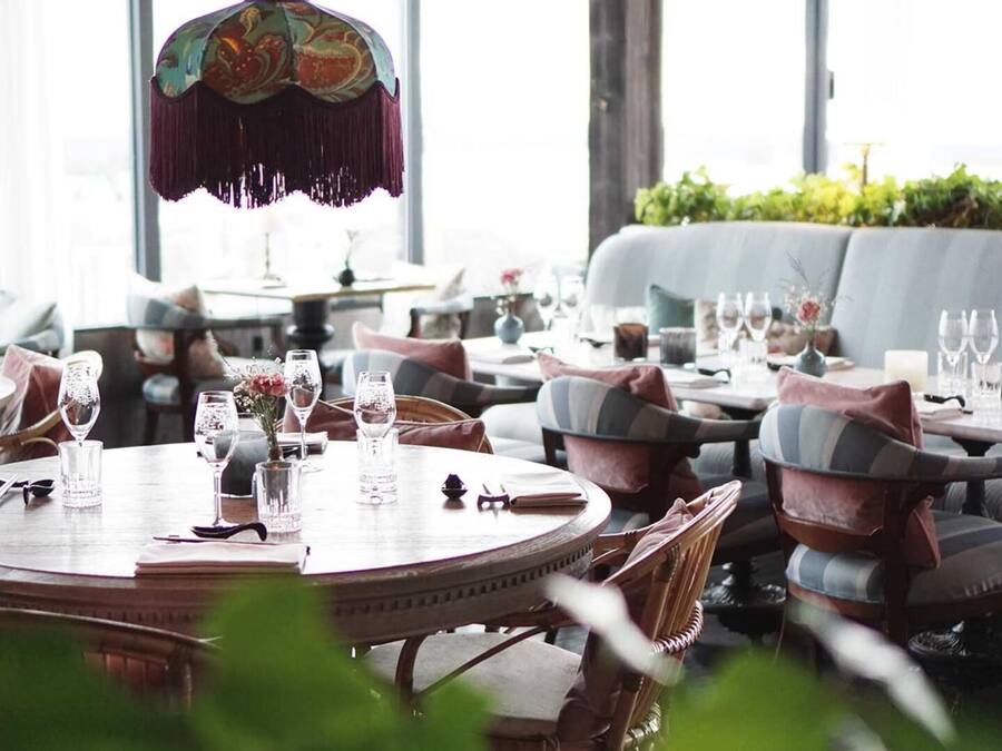 Un restaurante con mesas redondas y sillas acolchadas, ventanas amplias y una lámpara decorativa colgante.