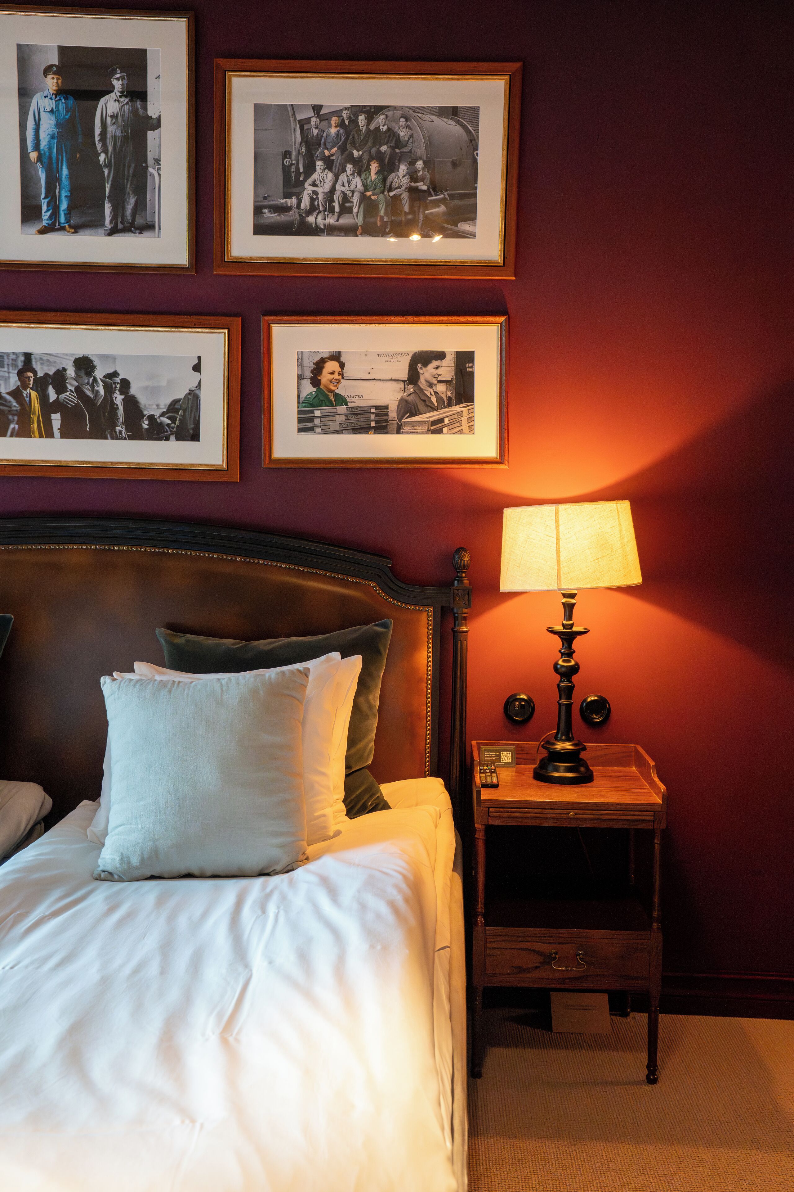 A hotel room with a neatly made bed, a bedside table with a lamp, and framed photographs on the wall.