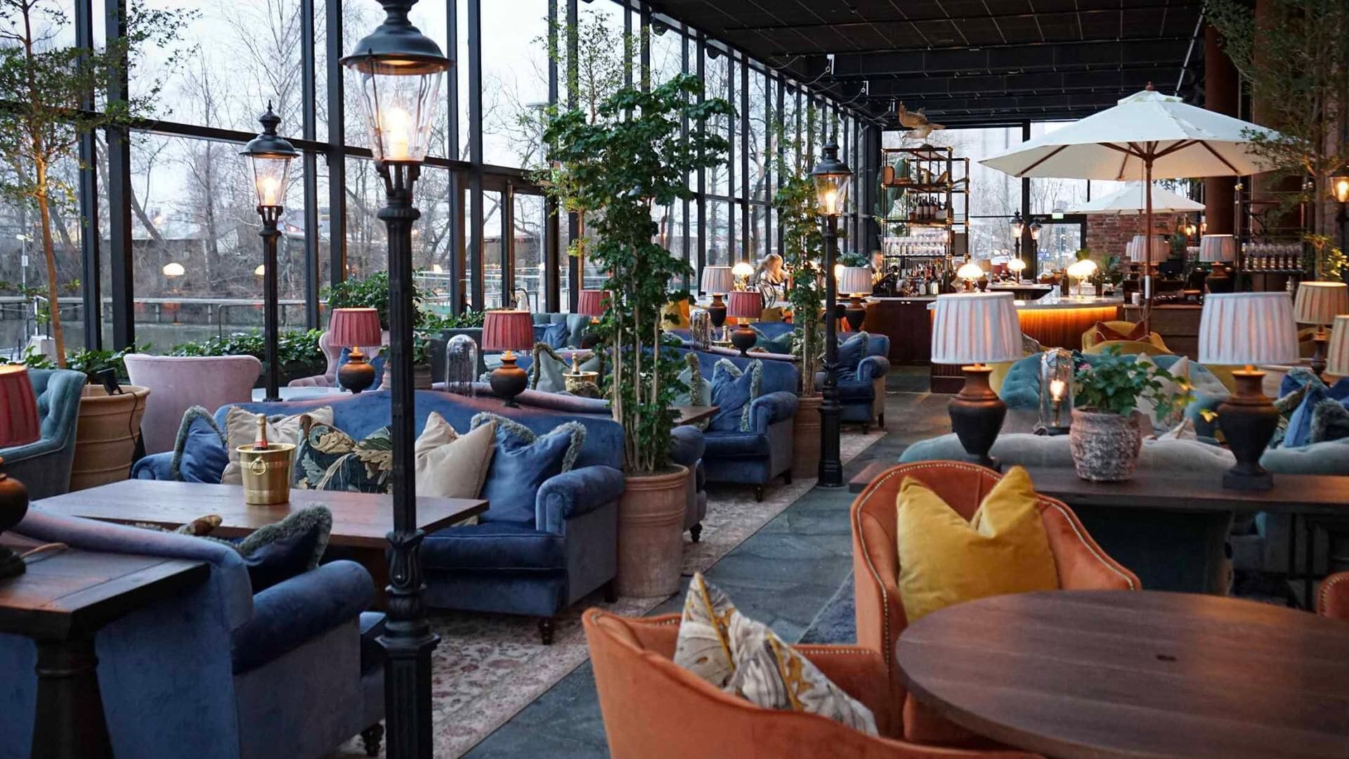 A cozy, well-lit outdoor seating area with plush sofas and tables, surrounded by large windows and potted plants.
