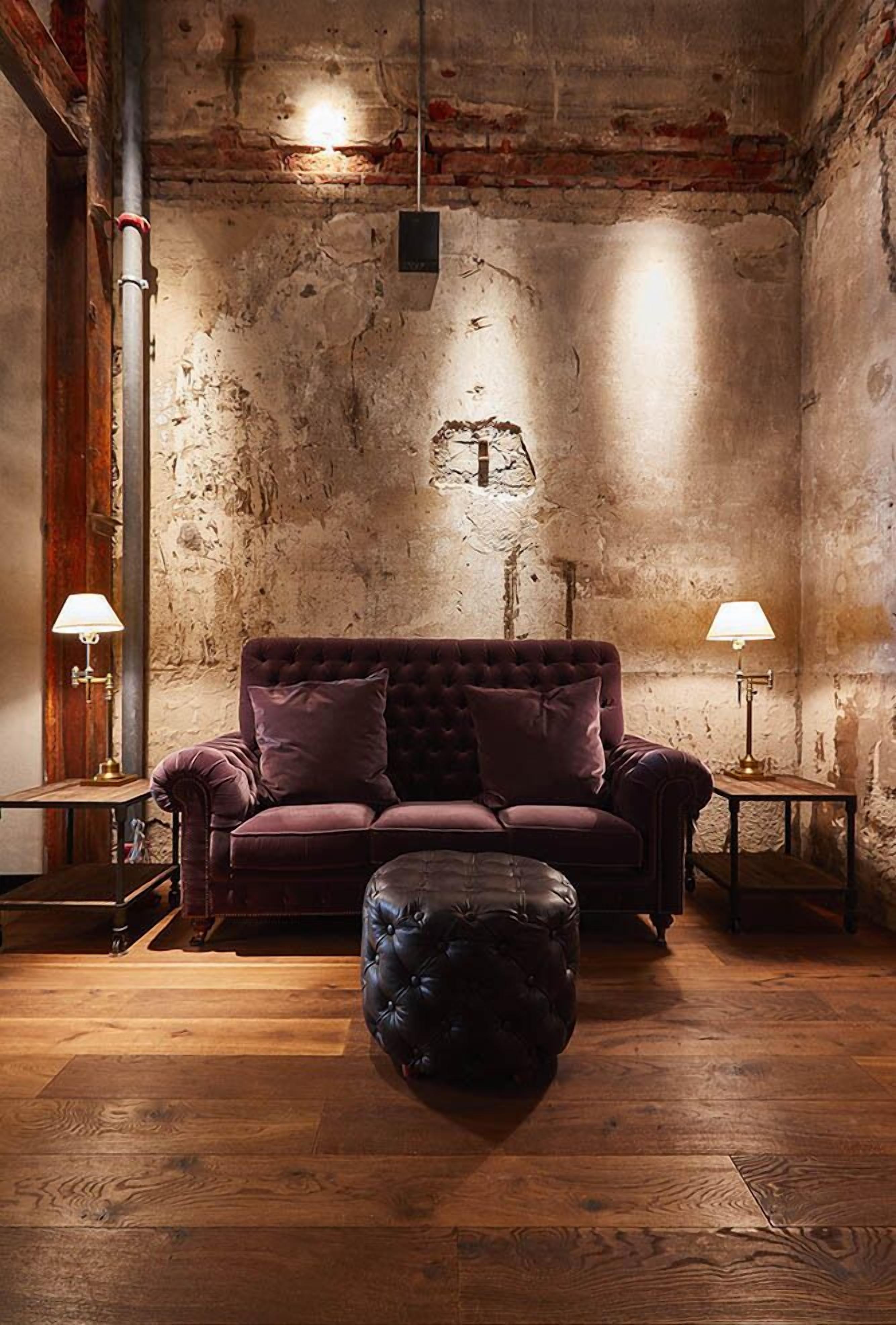 A room with a rustic brick wall, a tufted sofa with cushions, a leather ottoman, and two side tables with lamps.