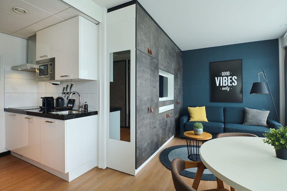 Een moderne keuken met witte kasten en een zwart aanrecht, een eethoek met een ronde tafel en stoelen, en een woonkamer met een blauwe bank en een bordje met de tekst 'Good Vibes Only'.