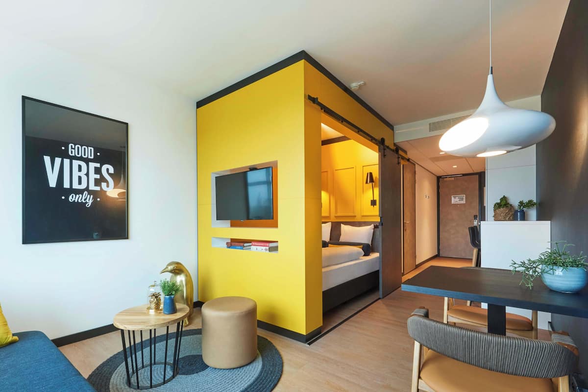 Een moderne hotelkamer met een geel accentmuur, een bed, een bureau en een zithoek.