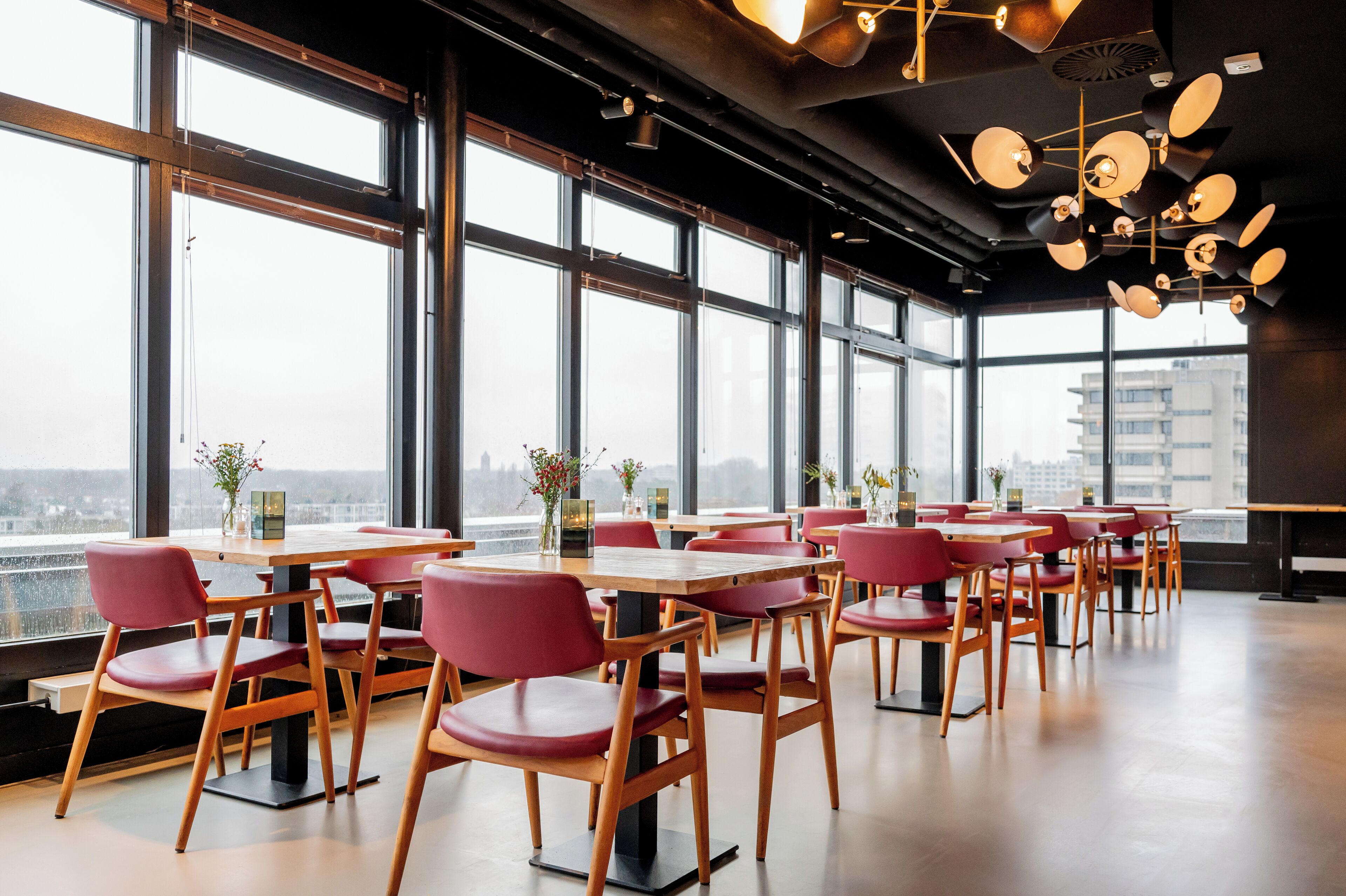 A modern restaurant with large windows, wooden tables, and red chairs.