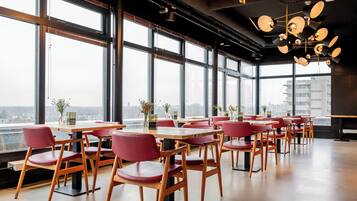 A modern restaurant with large windows, wooden tables, and red chairs.