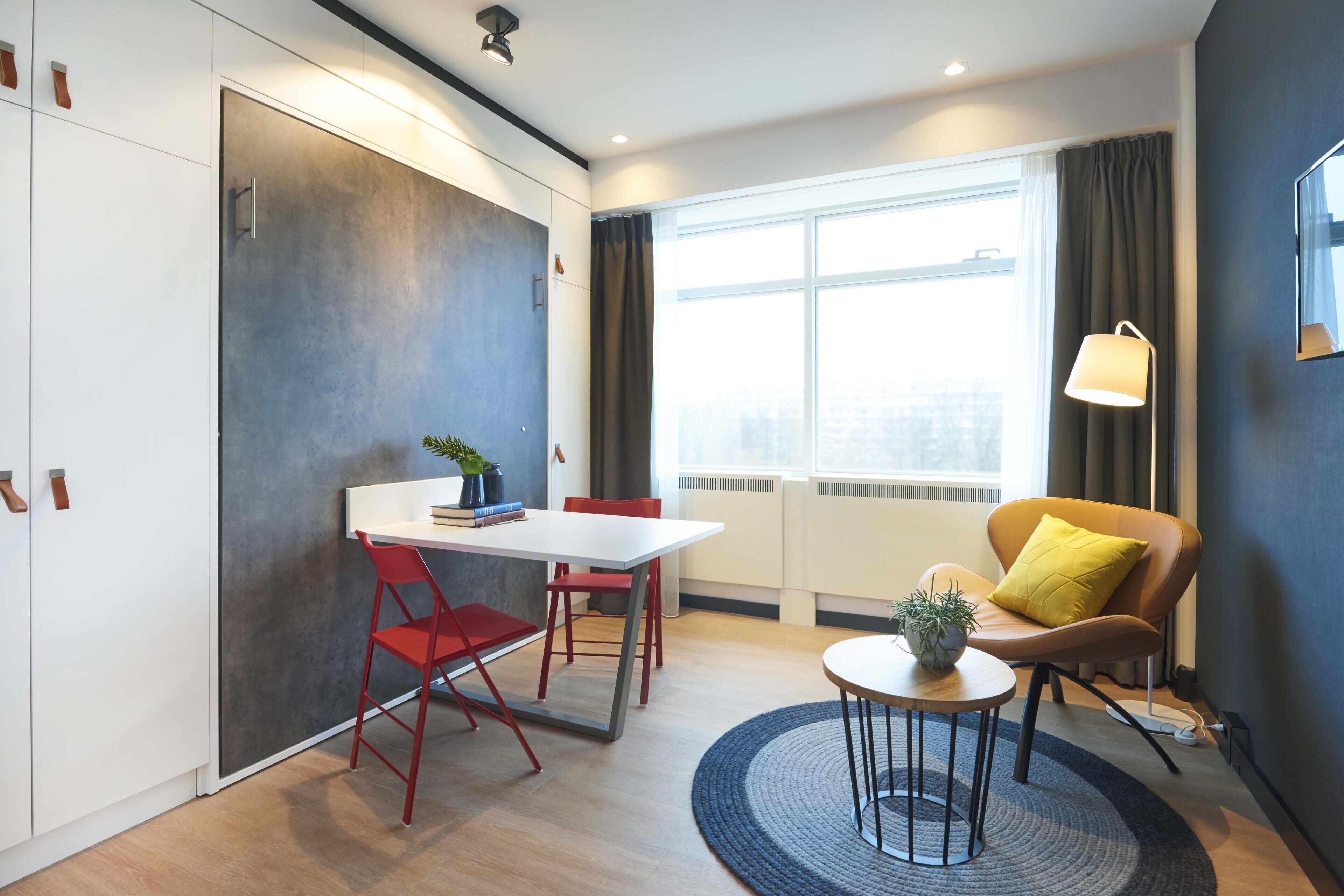 Une pièce moderne avec une table blanche, des chaises rouges, une table basse ronde, un coussin jaune et une lampe.