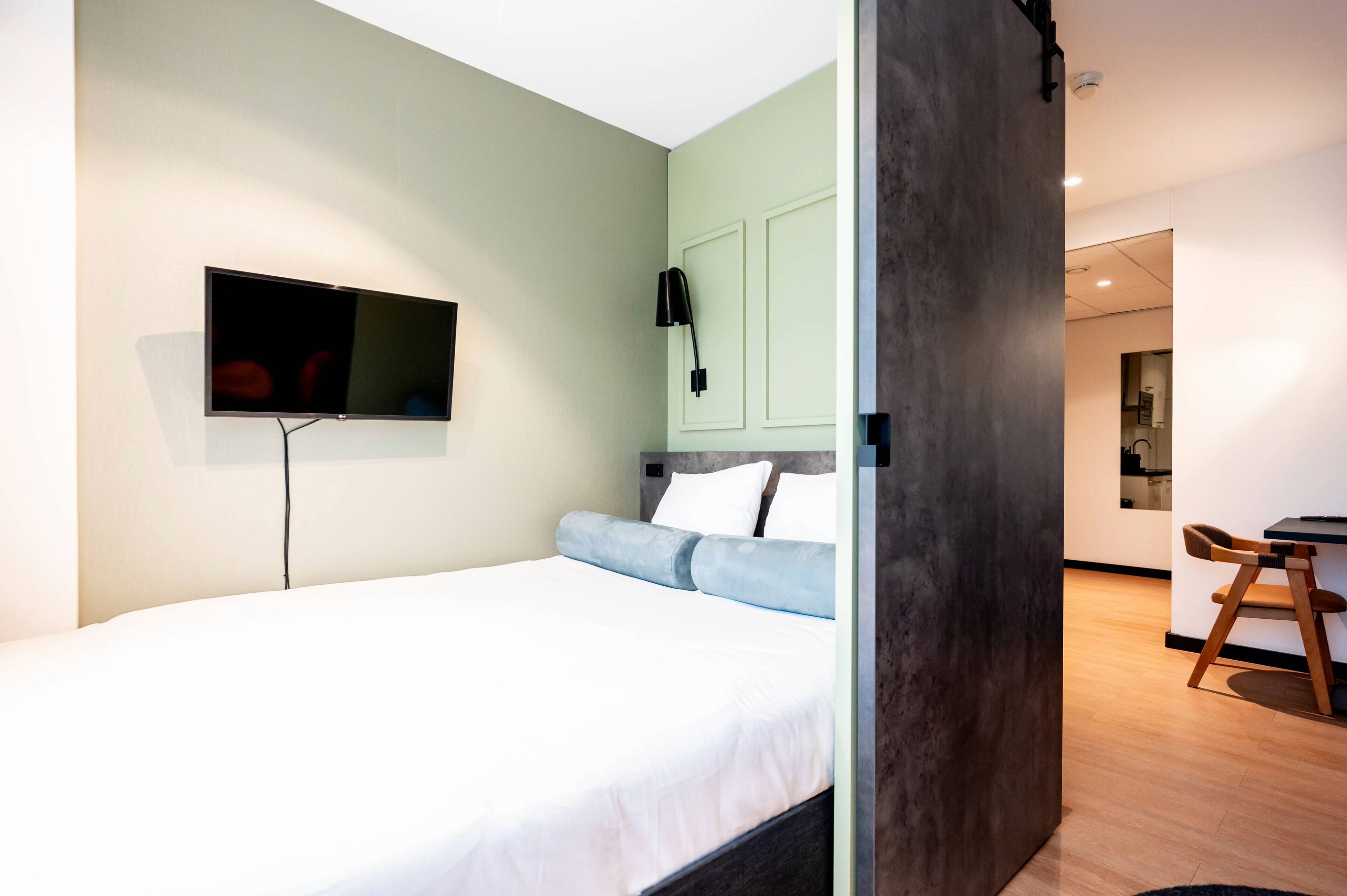 A modern bedroom with a bed, a TV mounted on the wall, and a desk with a chair in the adjacent room.