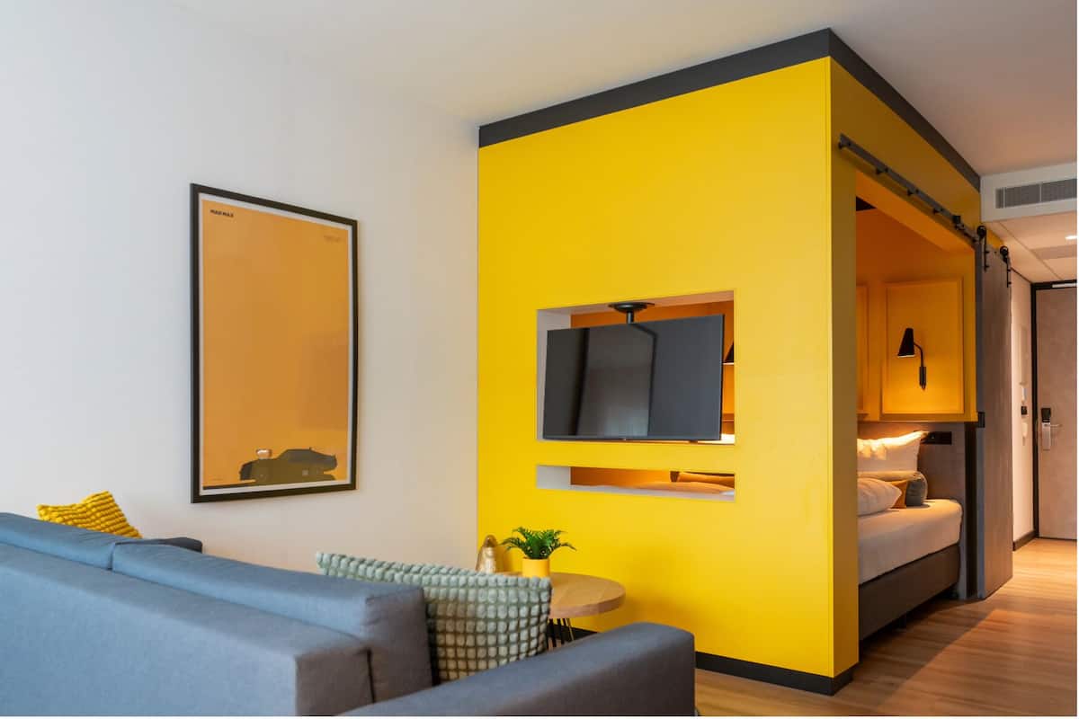 Een moderne hotelkamer met een heldergele accentmuur, een flatscreen televisie die eraan bevestigd is, een grijze bank, een klein rond tafeltje met een plant, en een bed met hoofdbord.