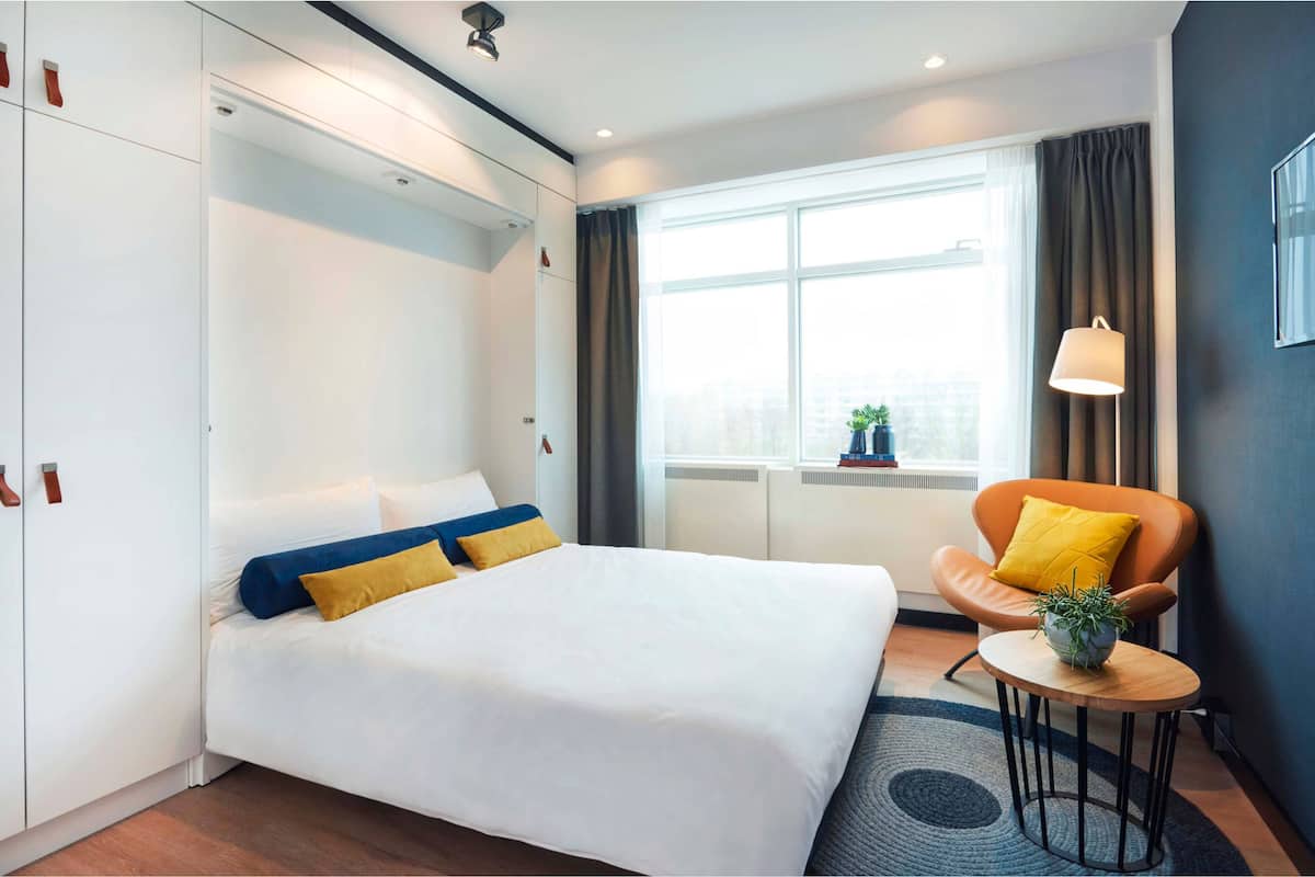 Een moderne hotelkamer met een groot bed, een klein rond tafeltje met een plant, een stoel met een geel kussen en een raam met gordijnen.