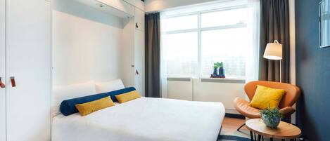 Una habitación de hotel moderna con una cama grande, una mesita redonda con una planta, una silla con cojín amarillo y una ventana con cortinas.