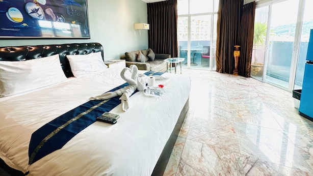 A hotel room with a neatly made bed, a sofa, a small table, and a view of the outside through large windows. - Koenig Hotel Pattaya (Pattaya)