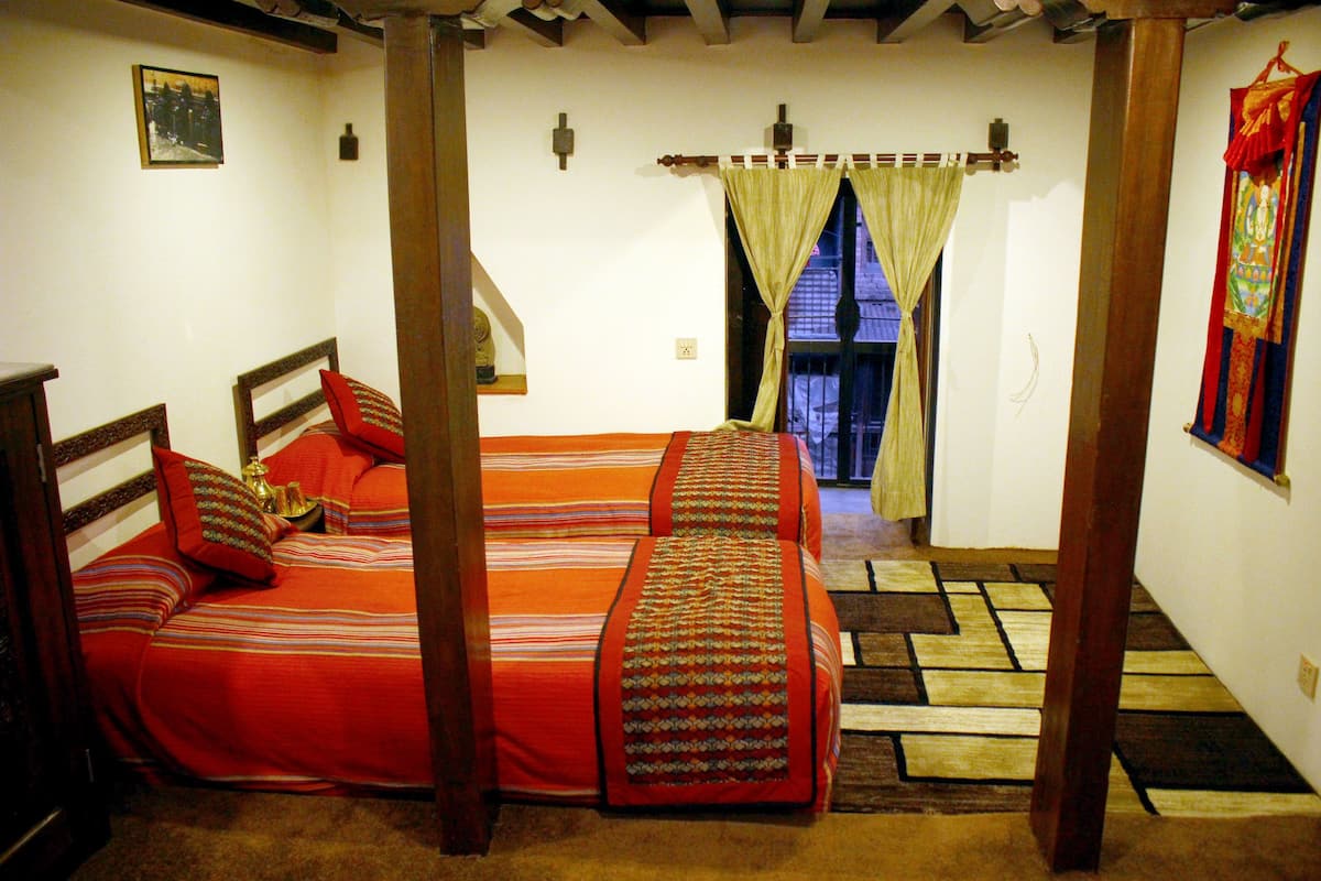 A room with two beds, a window with curtains, a wooden floor, and a wall-mounted light fixture.