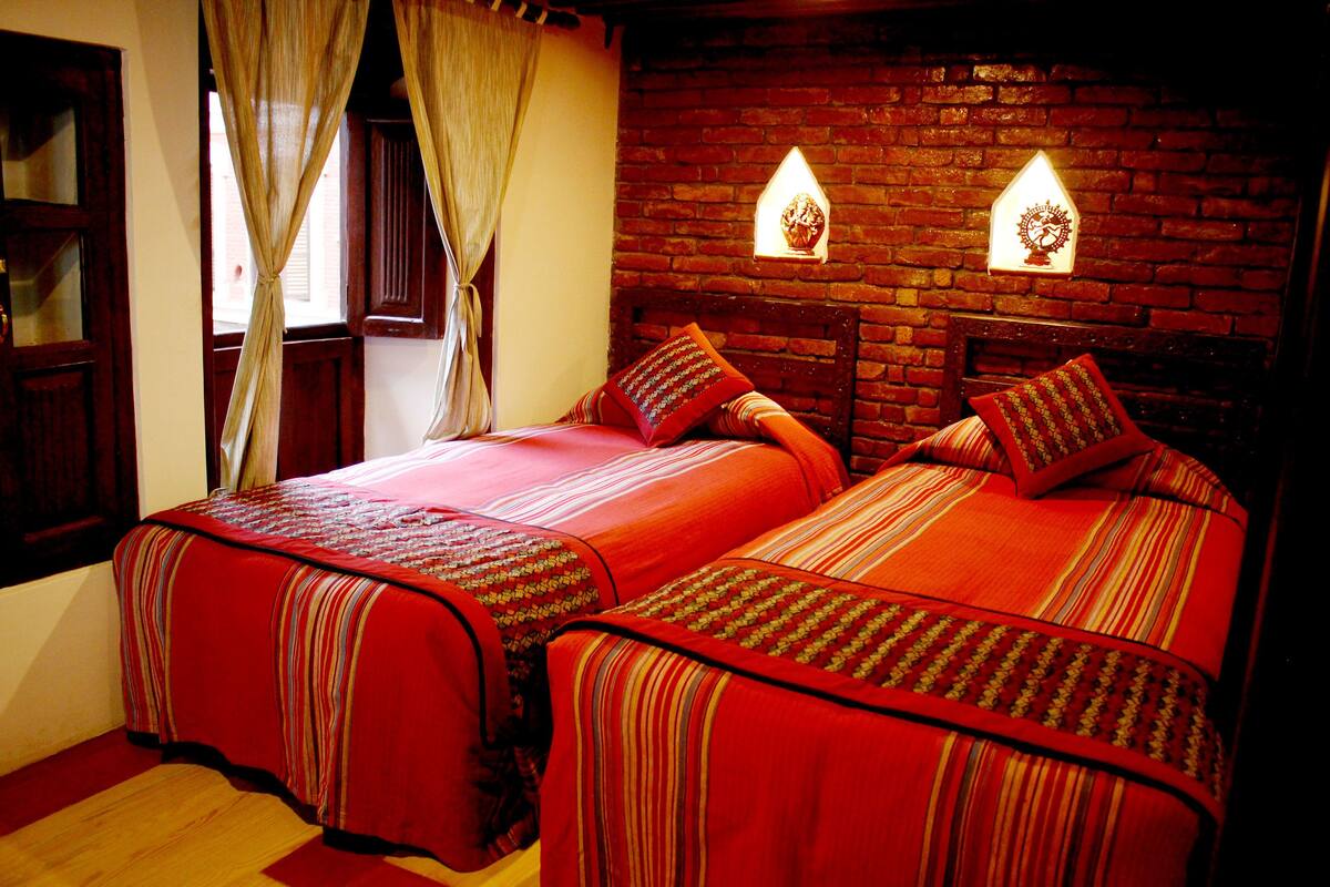 A room with two beds, each with red bedding and striped pillows. There are two wall-mounted light fixtures and a window with curtains.