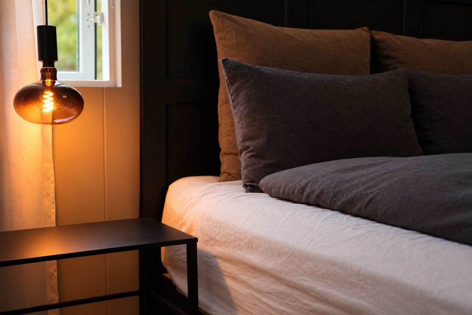 A bed with dark bedding and pillows, a bedside table, and a hanging light fixture.