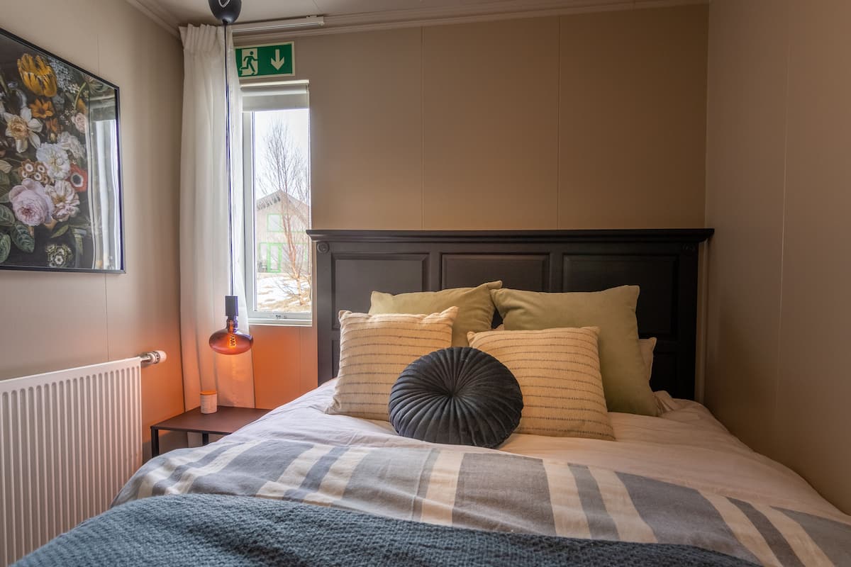 A neatly made bed with striped bedding and decorative pillows, a radiator, a framed floral artwork on the wall, and a window with a view of the outside.