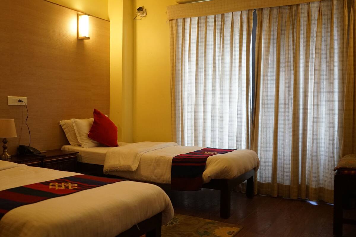 A hotel room with two beds, a nightstand with a lamp, a wall-mounted light, and a window with curtains.