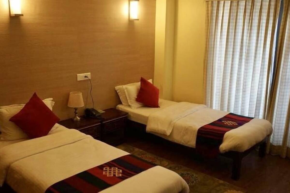 A hotel room with two single beds, each with a red pillow and a blanket with a logo. There is a nightstand with a lamp, a window with curtains, and a wooden wall.