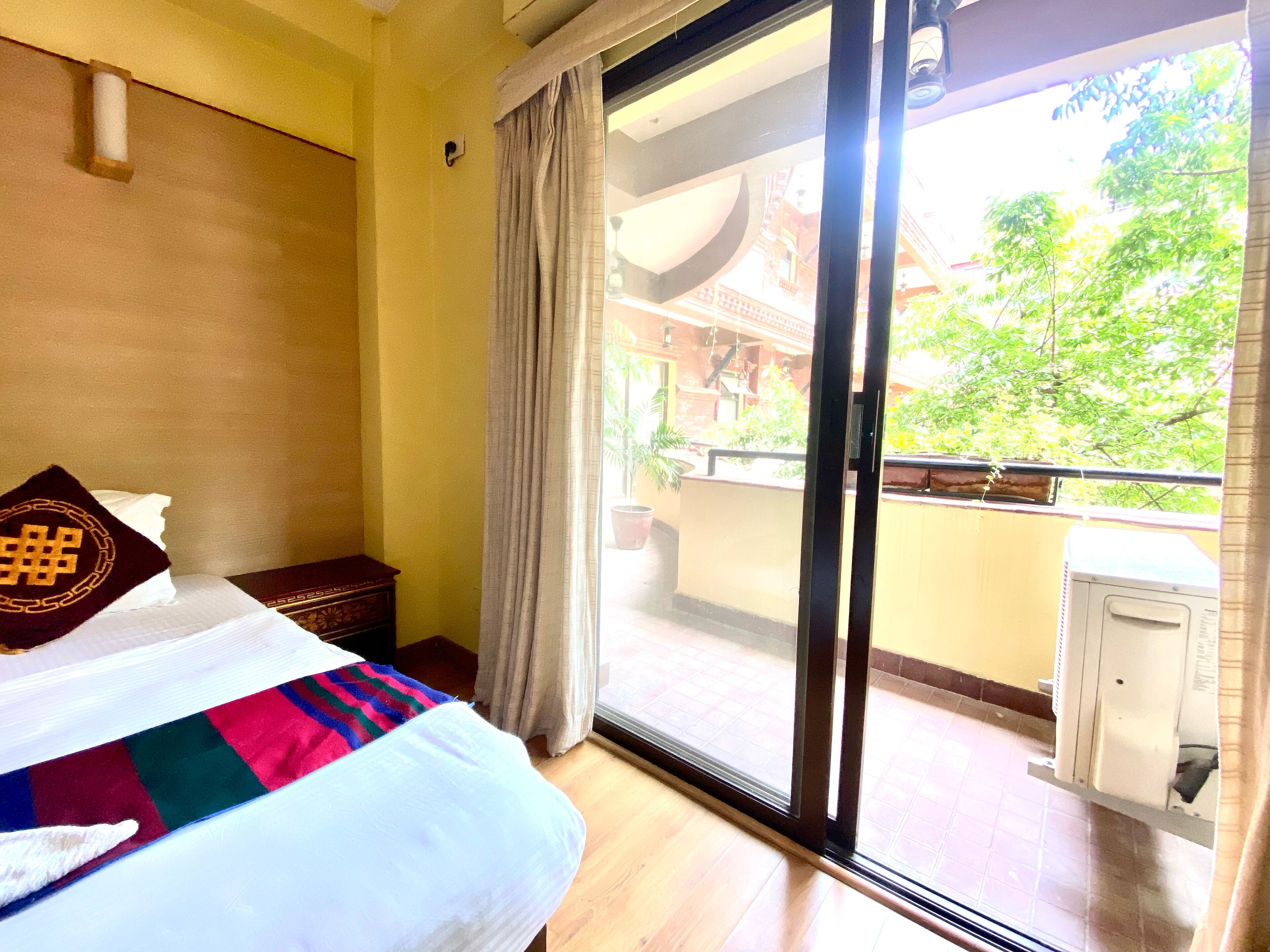 Habitación de hotel con cama, ventana con vista a un edificio y follaje, y puerta corrediza a un espacio exterior.