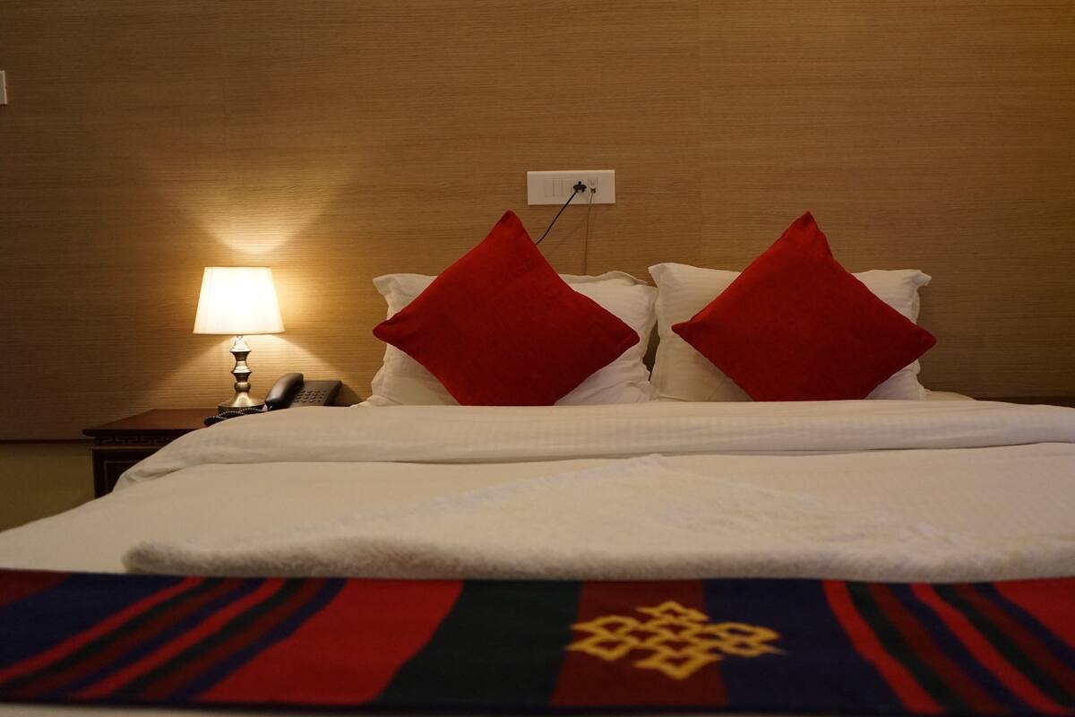 A hotel room with a neatly made bed, a bedside lamp, a phone, and a power outlet on the wall.