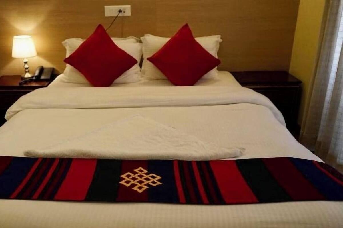 A hotel room with a neatly made bed, two red pillows, a white throw blanket, and a patterned bedspread.