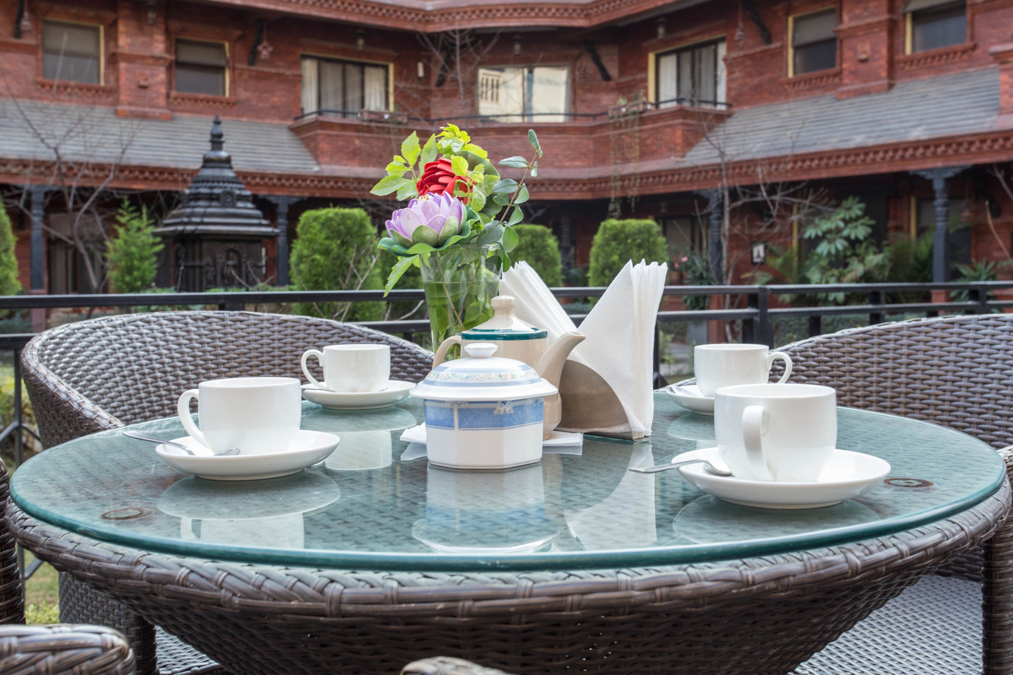 Una mesa redonda de vidrio con sillas de mimbre, tazas y platitos blancos, una tetera y un florero con flores en un patio de hotel.